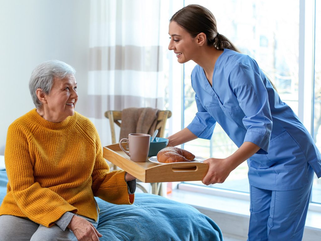 A caregiver bringing a tray of tea and a pastry to an older woman sitting by a window.