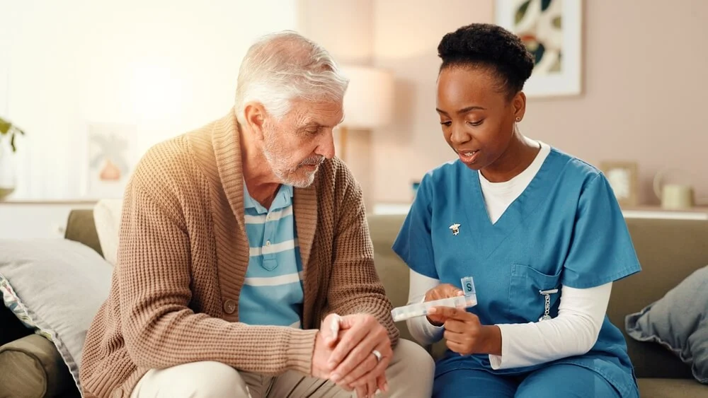 A caregiver and an older man looking together at a day-of-the-week pill organizer on the sofa.