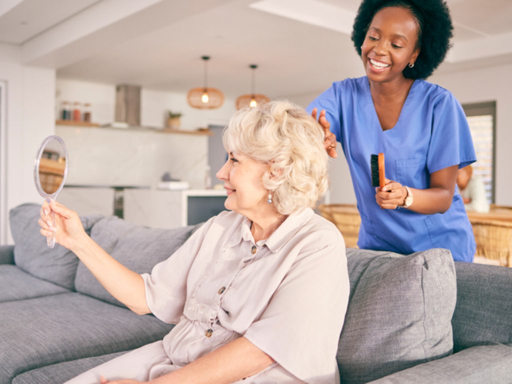 An older woman holding a hand mirror while a caregiver brushes her hair in her living room.