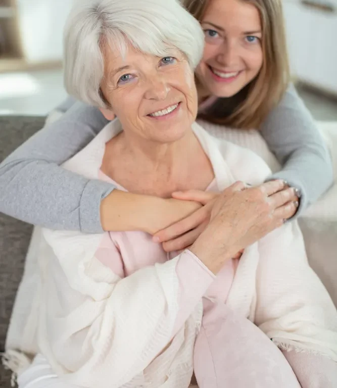 An older woman seated at home with a younger woman beside her, both smiling